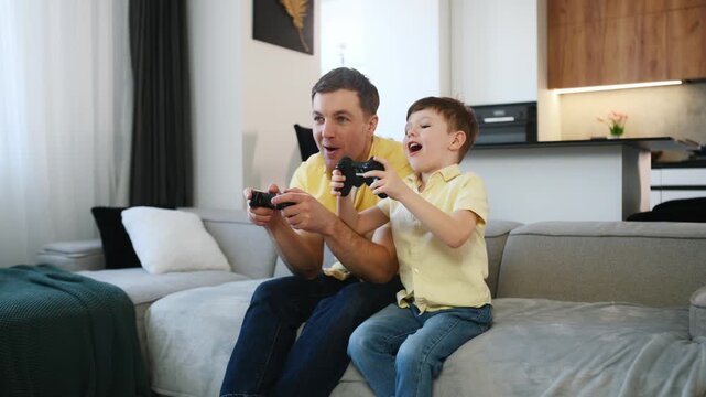 Father and young son enjoy playing video games on a sofa in a contemporary living room, with a kitchen area visible behind them