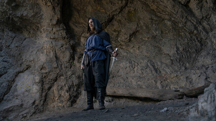 A lone, hooded figure stands before a rocky cliff face, clutching a sword and gazing into the shadows. © horimono