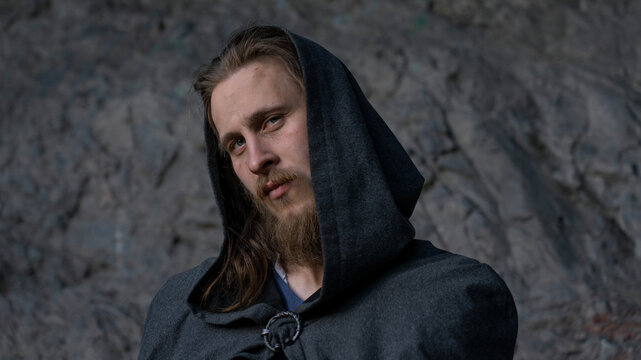 A man with a long beard and hooded cloak gazes thoughtfully against a textured stone wall.