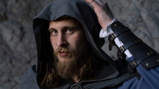Intense gaze of a bearded man with hood and leather bracers against textured stone backdrop.