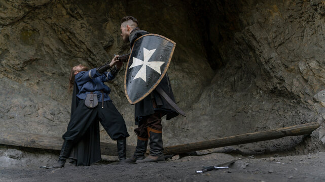 A knight with a shield defends against an attacker in a rocky, medieval setting.