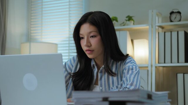 Asian Woman Writing Notes While Working on Laptop at Office Desk. Productive Professional Multitasking and Work Lifestyle.