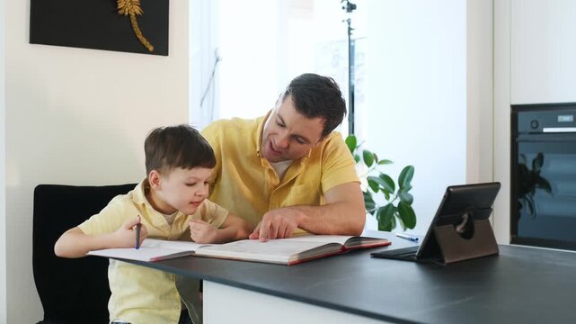 Father assists his young son with a learning activity at home, seated at a table with an open book and writing tools in a well-lit room