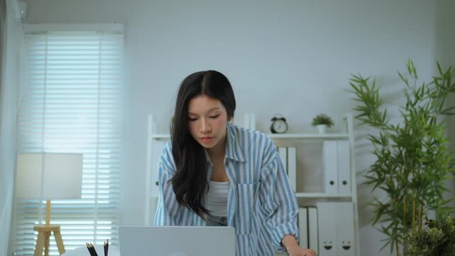 Asian Woman in Striped Shirt Standing and Holding Documents Looking Stressed in Office. Work Pressure and Overwhelmed Professional Lifestyle.