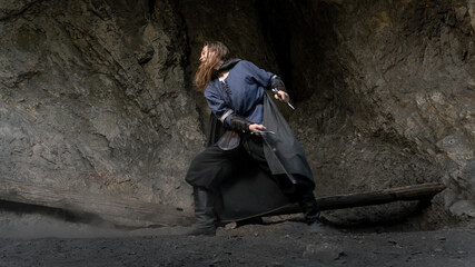 A rugged man with a long beard and dark clothing stands defiantly within a shadowy cave, clutching a sword. © horimono