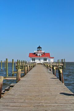 The Roanoke Marshes Lighthouse stands at the end of a long wooden pier extending into Roanoke Sound, framed by marina pilings and clear coastal skies in Manteo, North Carolina.