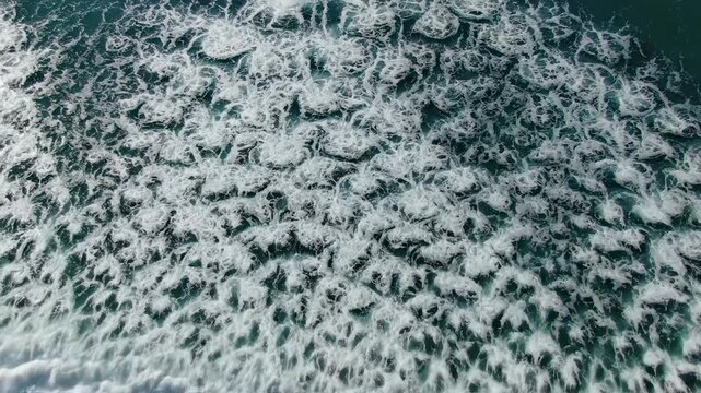 Natural patterns form as turbulent ocean waves crash over underwater reefs creating bubbling textures in the turquoise sea which captures the raw power of moving water during a sunny afternoon