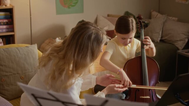 Medium long shot of Caucasian female tutor guiding kid playing cello during private music lesson at home. Footage is perfect for advertising music schools, tutoring services, and arts education