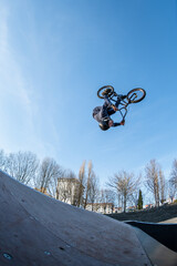 Naklejka premium Low angle shot of a young man doing an extreme flip stunt on a bicycle above a wooden ramp. The clear blue sky background emphasizes the high altitude and adrenaline of urban extreme sports.