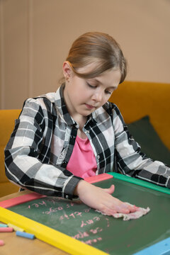 Girl cleaning a small colorful chalkboard on a wooden table, erasing pink chalk marks with a wet cloth, focusing on the task while developing her concentration and fine motor skills. Vertical photo