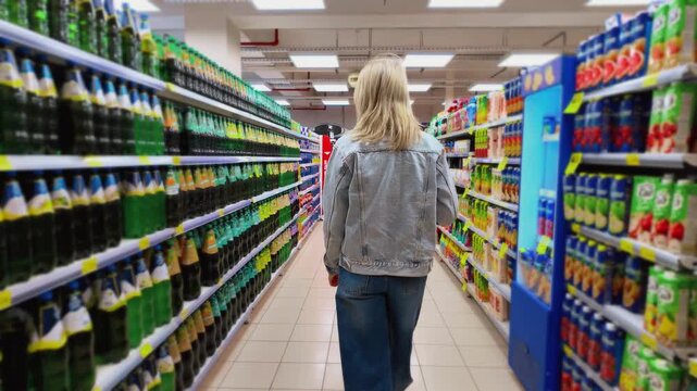 Rear view of a young woman in a denim jacket walking through the beverage aisle of a large grocery store, browsing the extensive selection of juices and sodas on the shelves