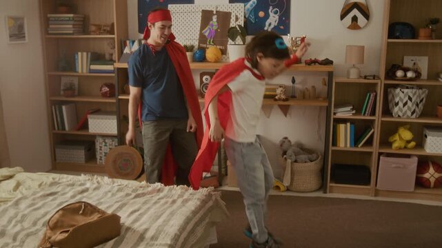 Young man and child wearing superhero capes playing active game, jumping on bed. Clip is perfect for family bonding concepts, parenting campaigns, or toy commercials