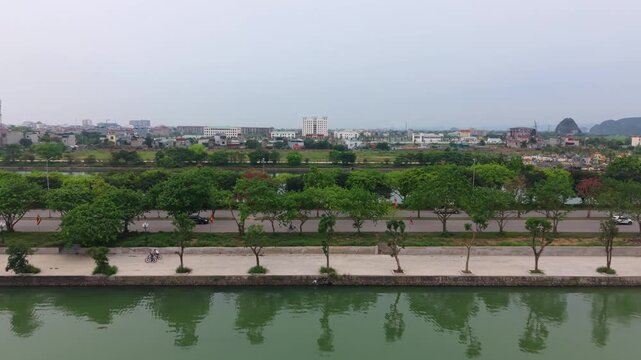 Lateral drone slider shot moving across a lakeside promenade in Ninh Binh featuring cyclists, tree-lined pathways, and reflections mirrored across calm water.