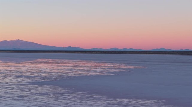 Salt flat landscape with reflective saline surface and distant Andean silhouettes at Salinas del Diamante, Mendoza, Argentina during twilight conditions, drone parallax orbit and yaw shot.