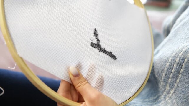 Hands creating cross stitch pattern on white fabric in wooden hoop with natural light