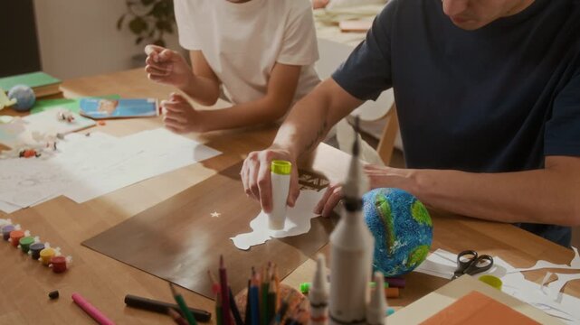 Asian father and multiethnic child gluing paper cutout onto environmental poster at wooden table. Clip is perfect for green energy promos, school projects, and ecology awareness campaigns