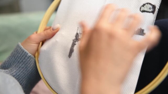 Person stitching black thread design on white fabric with embroidery hoop in cozy indoor setting