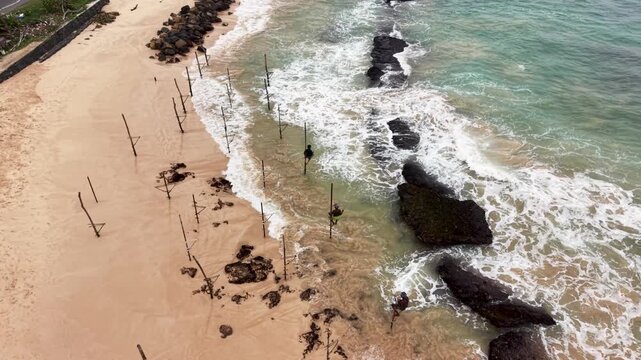 Aerial view of traditional stilt fishing poles with fishermen on a sandy beach with waves crashing against rocks in Koggala, Southern Province, Sri Lanka.