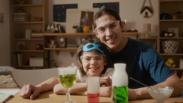 Portrait of young man and child wearing safety goggles, smiling at camera during home science experiment. Footage ideal for promoting STEM education and e-learning