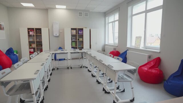 Classroom displays desks arranged in large semi-circle formation with colorful bean bags. Large windows fill interior with bright daylight