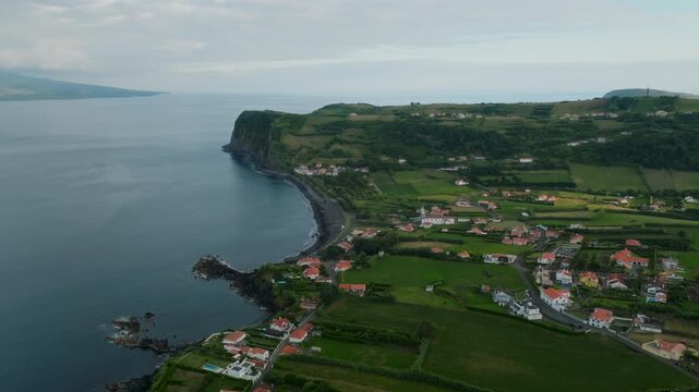 Aerial view of a coastal village on Faial Island in the Azores, featuring white houses with red roofs nestled between lush green fields, a dark volcanic sand beach, and the expansive Atlantic Ocean