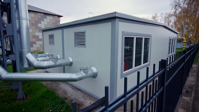 Large silver industrial pipes route directly from side of light grey facility structure up. Manicured lawn area adjacent to main building