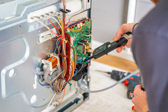 Technician testing electronic components and wiring inside disassembled electric oven. Appliance diagnostics and professional repair service concept