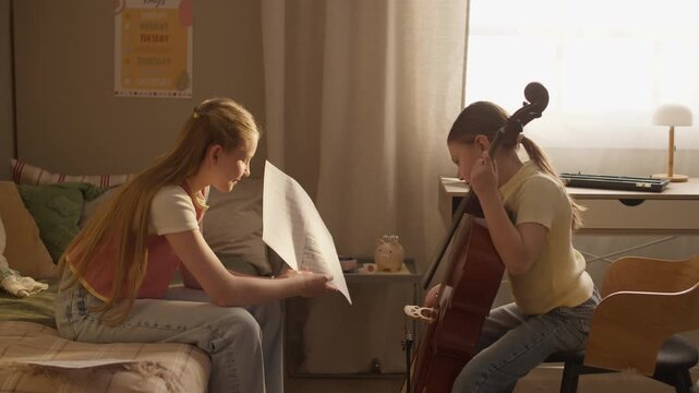 Side view of teenager holding sheet music and instructing child playing cello during home music lesson. Footage is ideal for promoting private tutoring, music schools, or creative education