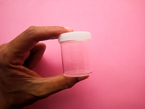 Hand holding an empty transparent medical specimen collection container with a white cap, isolated on a pink background.
