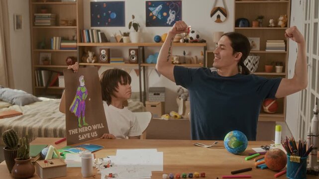 Father flexing muscles while young son holds superhero drawing at desk. Ideal for fatherhood campaigns, parenting content, and family lifestyle marketing
