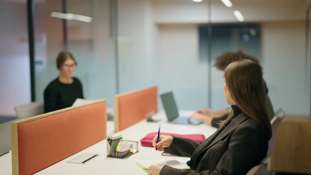 Businesspeople are engaged in focused work at their desks within a contemporary open-plan office. Coworkers concentrate on tasks, utilizing laptops and writing notes in a productive setting