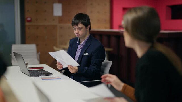 A male employee with Down syndrome works with documents at a bright office desk. He reviews papers received from a coworker while also using his laptop