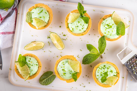 Mini Mojito Tarts, portioned Key Lime Pie tartlets cakes decorated with lime slices, mint and citrus zest, on white marble table background copy space
