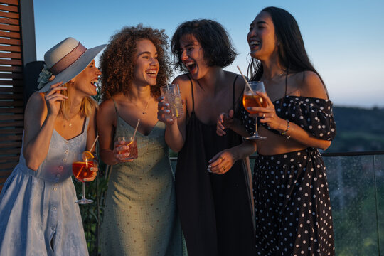 Young women friends laughing enjoying cocktails at sunset rooftop party