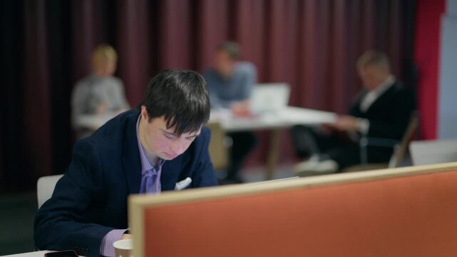 A young employee with Down syndrome focuses intently on his laptop in a modern office. Other colleagues are also working diligently in the collaborative workspace