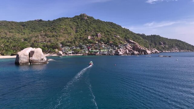 Drone view of a boat entering a tropical bay with blue water and rocky shoreline in Koh Tao, Thailand