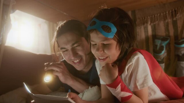 Young father holding flashlight and kid wearing superhero costume reading book in blanket fort. Footage suitable for family bonding and parenting