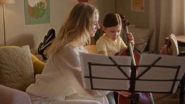 Arc shot of Caucasian female music teacher guiding young child playing cello during home lesson. Footage perfect for marketing private music tutoring, childhood education, or arts programs