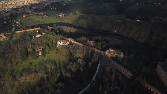 Aerial view of the historic Portico di San Luca winding through the lush green hills and residential villas under soft evening light in Bologna, Emilia-Romagna, Italy.