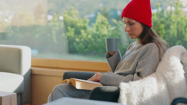 A woman reading by a large window and enjoying scenic snowy mountain view.