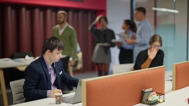 Young employee with Down syndrome works diligently on his laptop at an office desk. Various colleagues collaborate and focus on their tasks in a modern, inclusive workspace