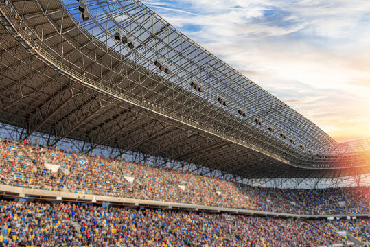 A large crowd of people fills the stands of a modern sports stadium under a massive steel roof structure at sunset.