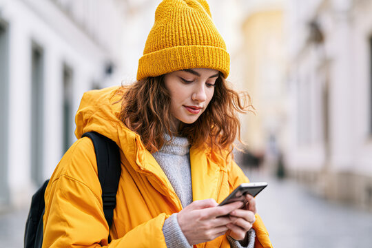 Young woman in a yellow jacket and beanie uses a smartphone outdoors in an urban setting, carrying a black backpack.
