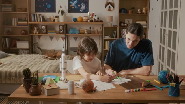 Young Asian father helping multiethnic child draw space rocket on paper at desk. Perfect for parenting, homeschooling, and creative STEM education campaigns