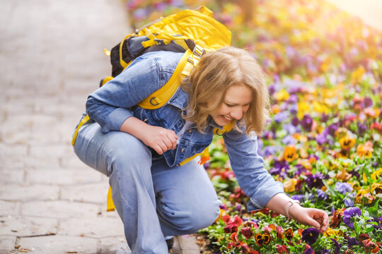 Smiling mid adult woman in denim jacket crouching by flower bed with pansies. Flower garden care and summer vacation concept.