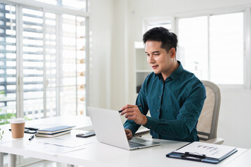 Fototapeta premium Young asian professional business man working on laptop, remote working using computer looking at screen watching seminar or searching information at office.