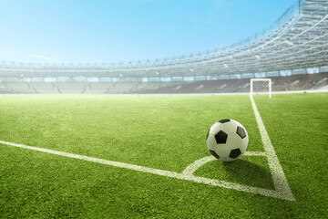 A black and white soccer ball on the corner kick line of a green field inside a large, empty...