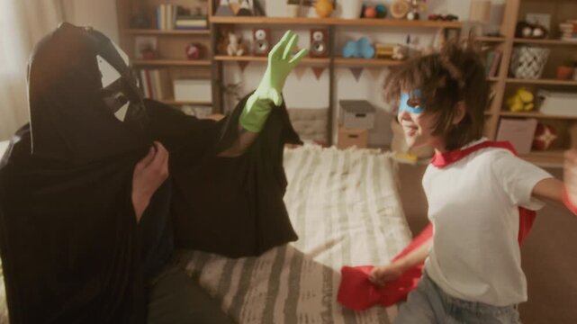 Zoom out shot of father wearing villain costume playfully fighting with kid dressed as superhero in bedroom. Clip suitable for parenting, family bonding, and Fathers Day marketing