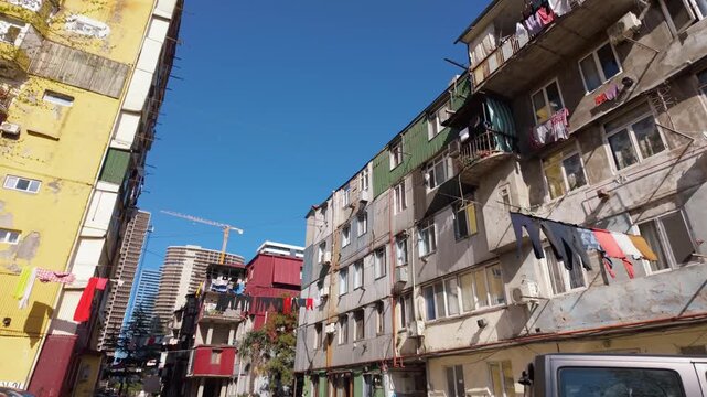 Low-angle view of laundry drying on clotheslines strung between old, weathered apartment buildings in a poor neighborhood, showing a stark contrast with modern skyscrapers in the distance