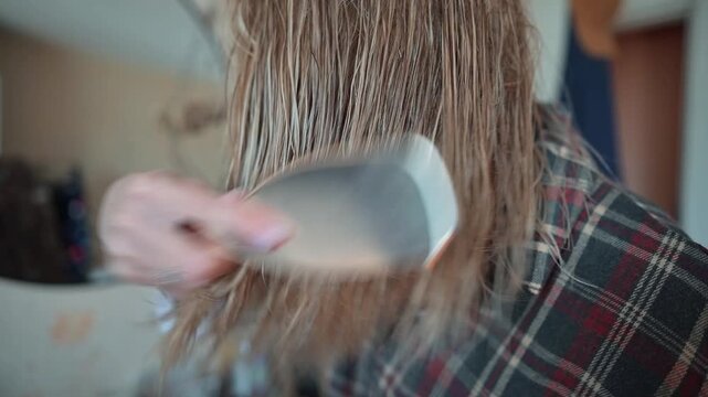 A young woman with long hair is brushing her wet hair . She demonstrates proper brushing technique with consistent strokes while looking into a mirror